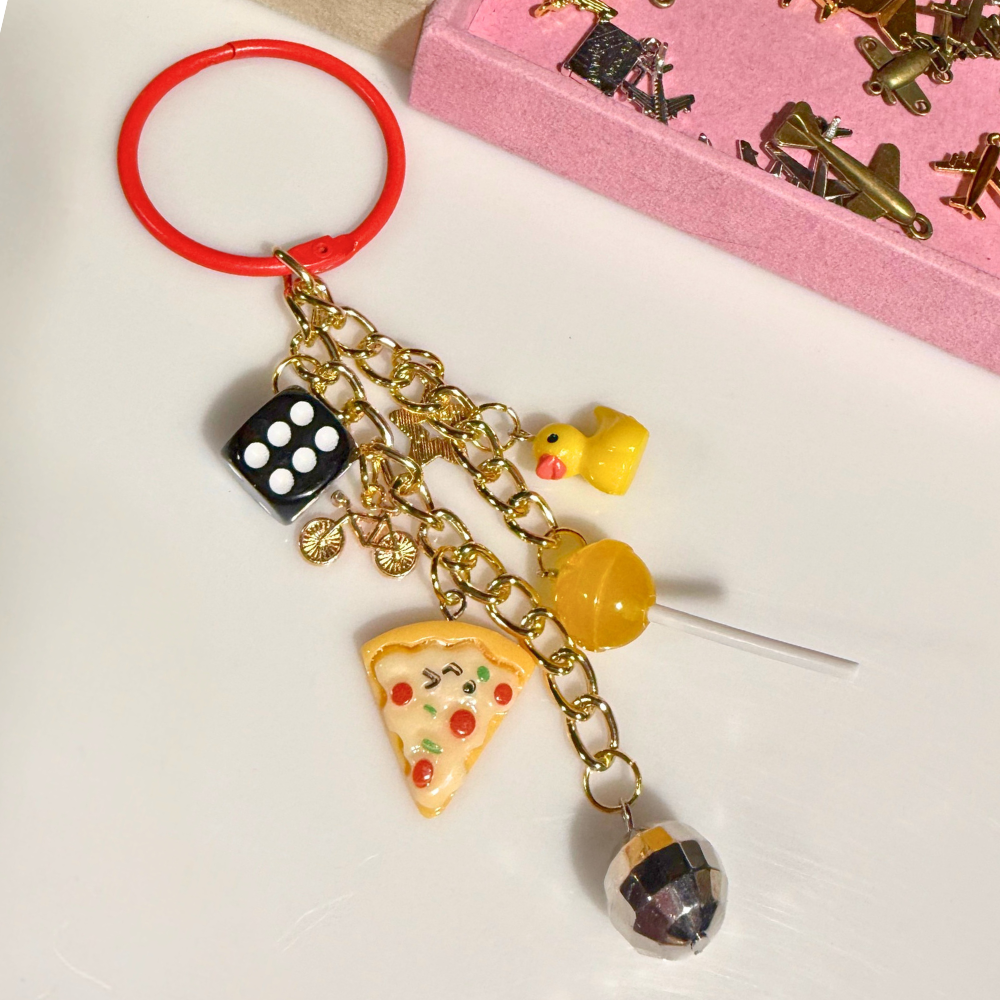 Keychain with various charms including a pizza slice, dice, and bottle cap on a white background.