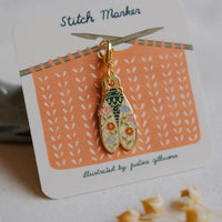 A gold-tone enamel stitch marker charm shaped like a cicada with floral details, mounted on a white display card labeled 'Stitch Marker' with an orange knitted pattern illustration.