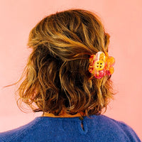 Wavy brown hair is secured with a large, yellow and pink daisy-shaped clip featuring a button center against a soft pink background.