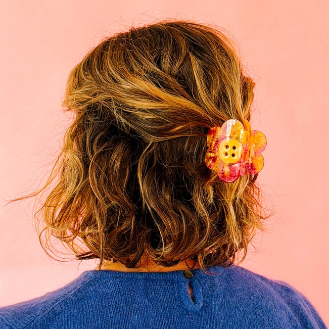 Wavy brown hair is secured with a large, yellow and pink daisy-shaped clip featuring a button center against a soft pink background.