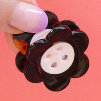A hand with a purple-tipped nail holds a flower-shaped claw clip with a tortoise border and white  button center against a coral background. 