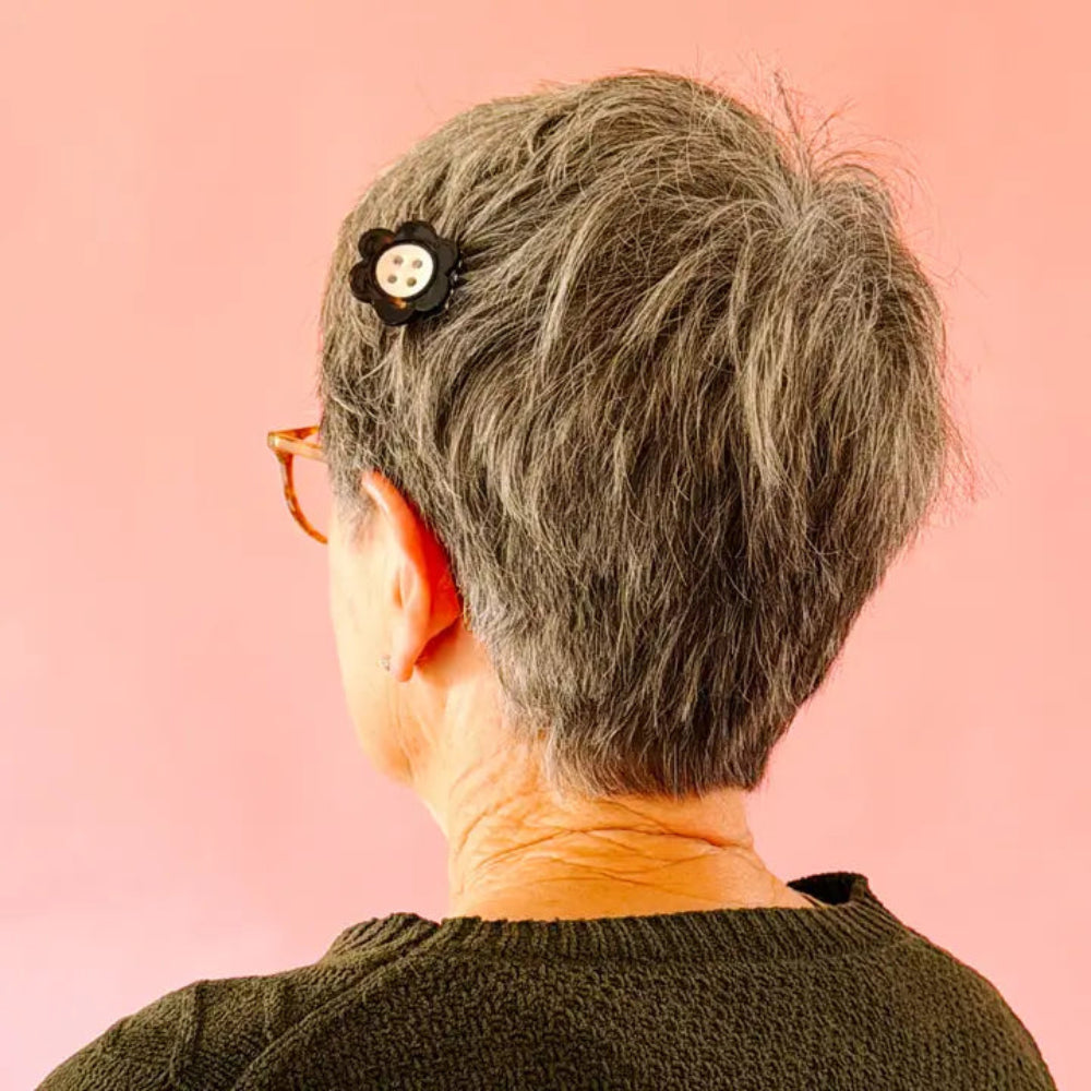 A person with short grey hair sports a mini daisy shaped claw clip against a pink background