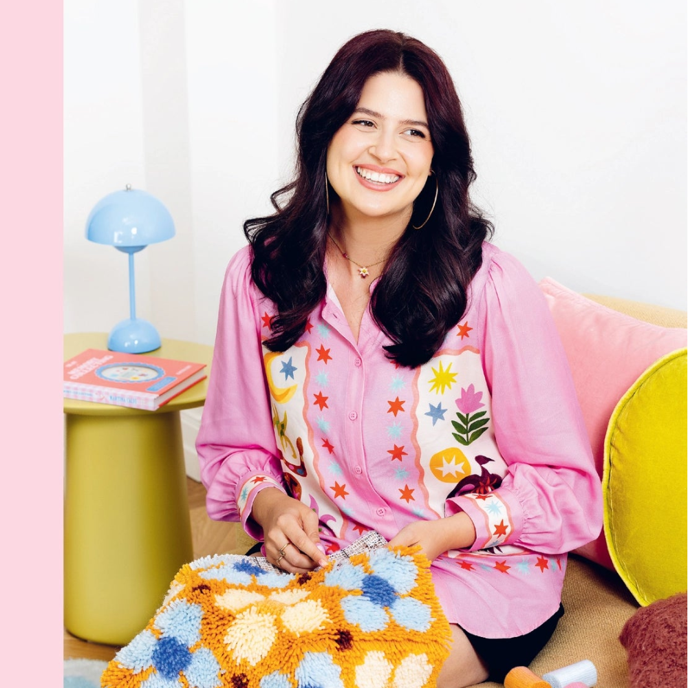 The author of Craft Club Yarn pictured seated on a couch working on a latch hook project from her book