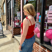 A profile view of a blonde woman wearing a red crochet tank top, standing in front of a storefront window.