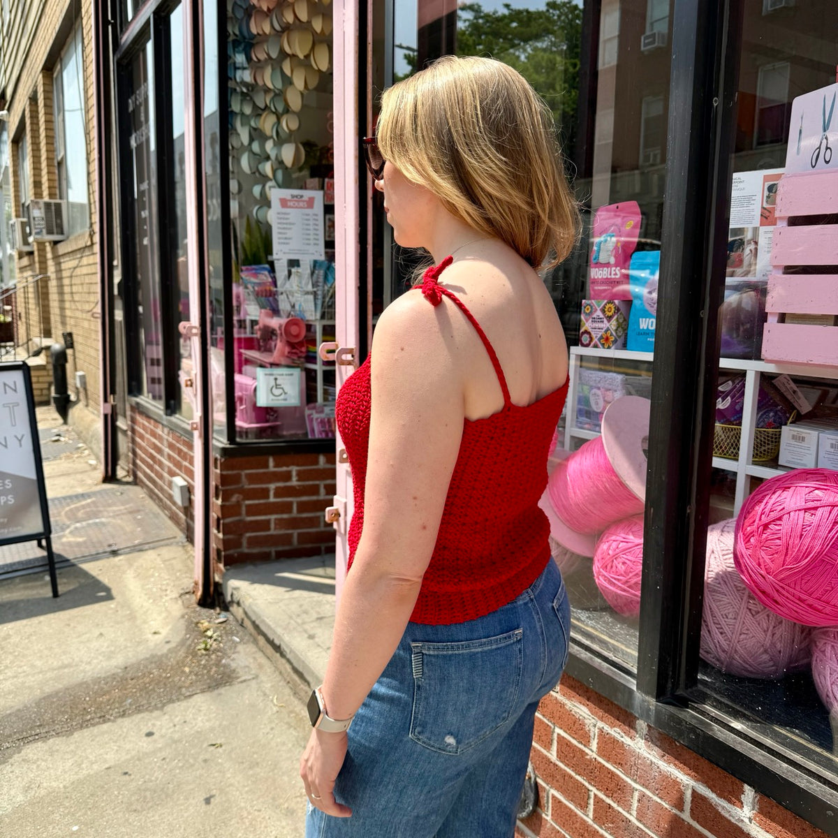 A profile view of a blonde woman wearing a red crochet tank top, standing in front of a storefront window.