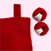 A close up of a red crochet tank top on a pink background, with two balls of cotton yarn.