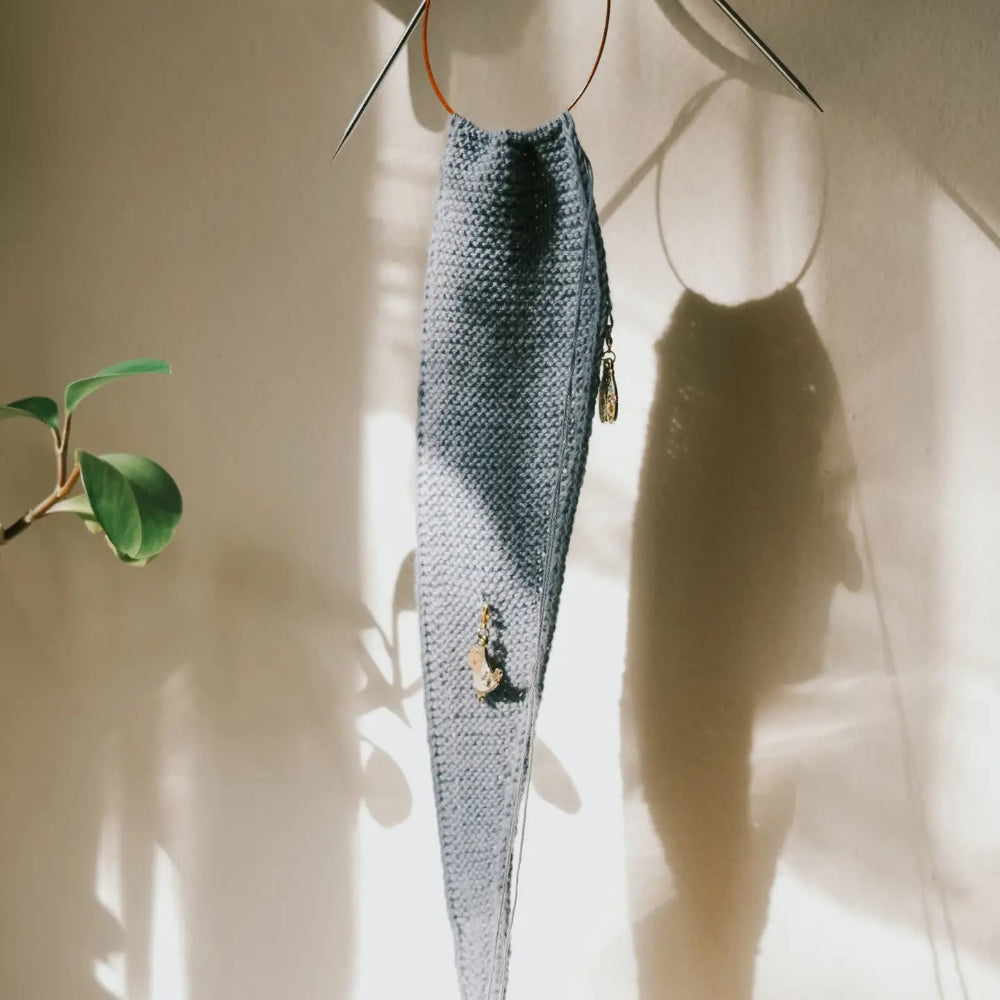 A long, narrow knitted scarf in a muted blue-gray color hangs from metal circular hooks against a warm beige wall, with sunlight casting shadows and a plant leaf visible on the left.