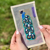 Needlepoint peacock design on fabric held by a person with a blurred green background