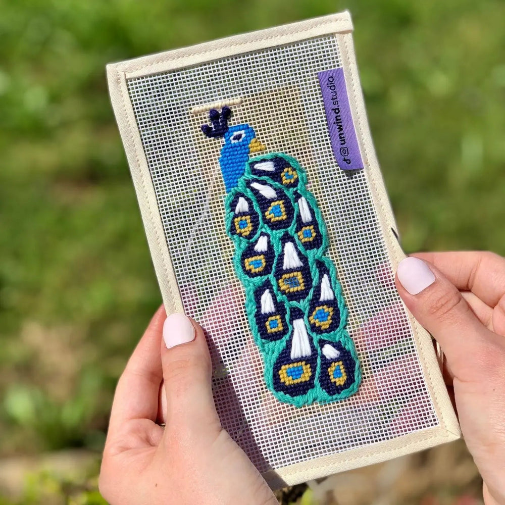 Needlepoint peacock design on fabric held by a person with a blurred green background