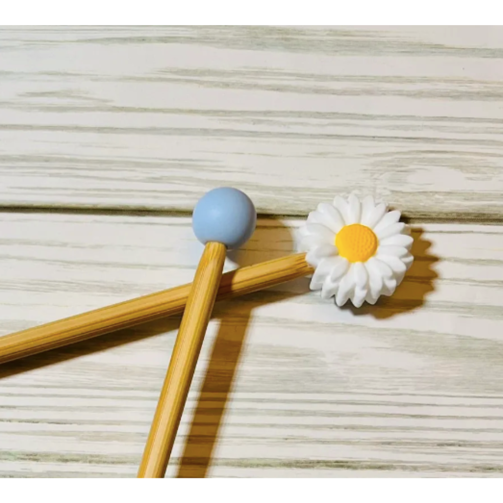 Two wooden knitting needles with colorful ends, one blue, and one daisy, on a wooden surface