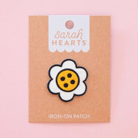 A Daisy-shaped iron-on patch with a yellow center and button dots is displayed on a brown card on a pink background