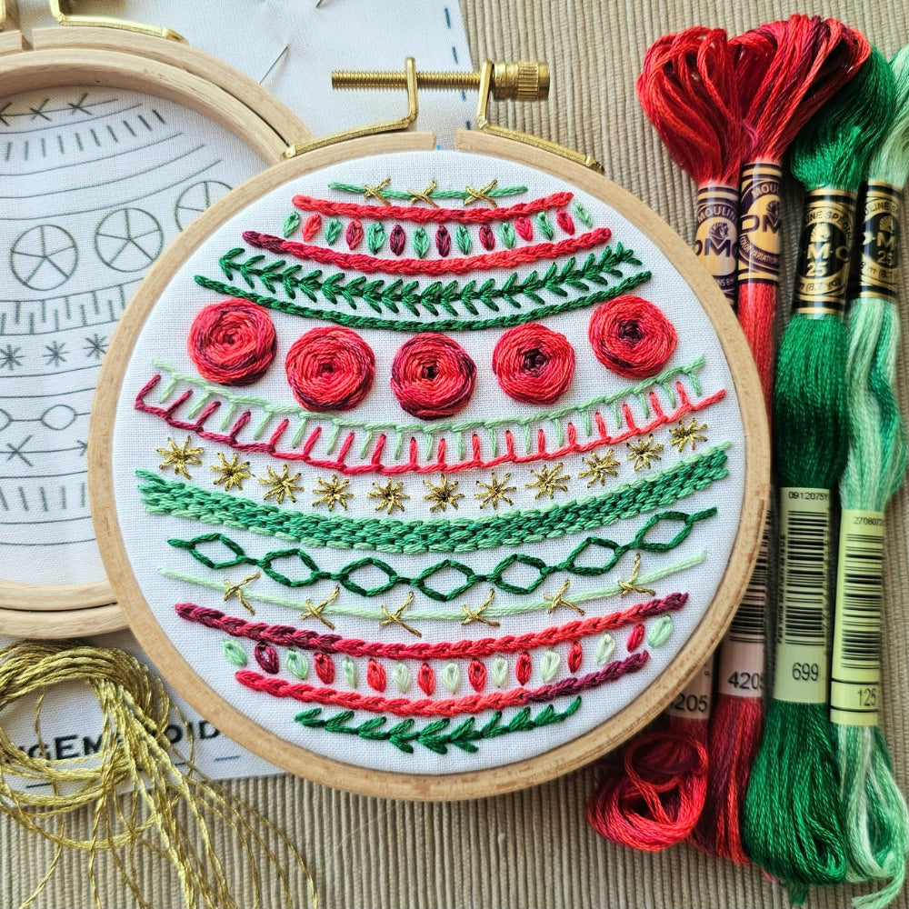 The "Deck the Halls" embroidery kit, showing an embroidery hoop with geometric stitching in red and green floss on a white background