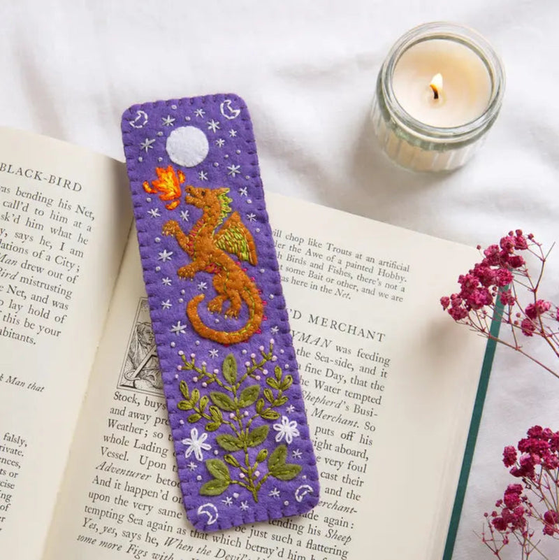 Embroidered bookmark with a dragon design on an open book next to a candle and flowers.