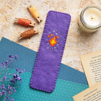Purple felt bookmark with embroidered  flame design on a textured surface with candles and spools of thread.