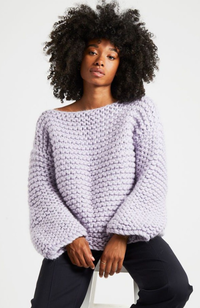 Woman with curly black hair wearing navy pants and a light purple oversized knit sweater.