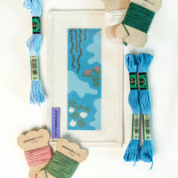 Needlepoint materials including thread, a hoop, and a partially completed design on a white background.