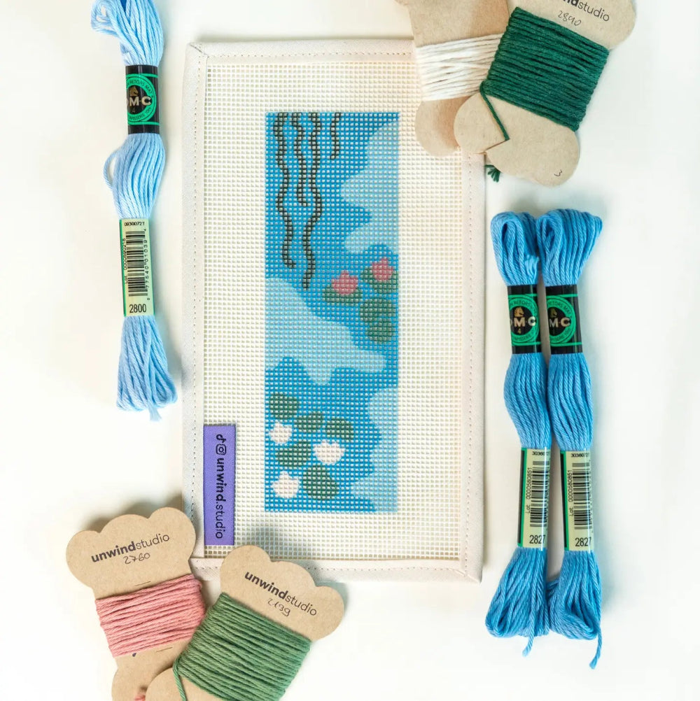 Needlepoint materials including thread, a hoop, and a partially completed design on a white background.