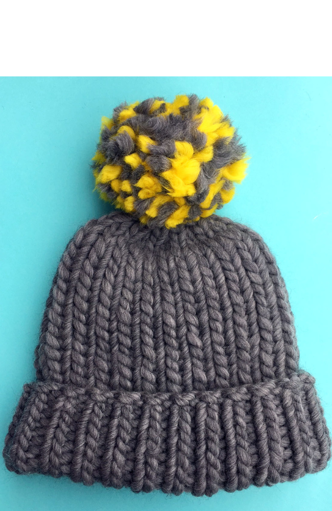 Gray knit hat with a gray and yellow pom pom laying flat on a bright blue background.
