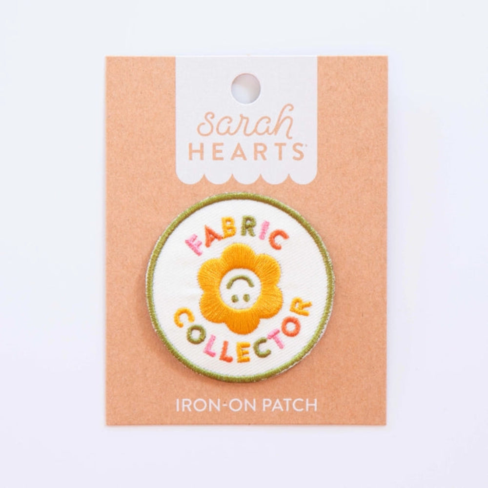 An iron-on patch featuring a smiling yellow flower with the words "Fabric Collector" in colorful letters on Sarah Hearts backing. 