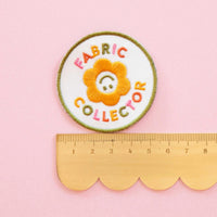 An iron-on patch with a yellow flower with the words "Fabric Collector" in colorful letters is on top of a scalloped ruler against a pink background. 