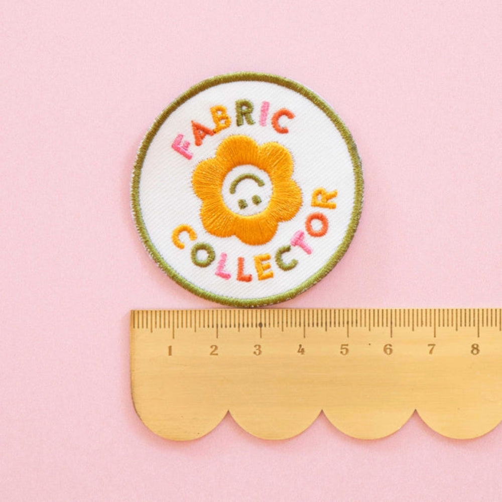 An iron-on patch with a yellow flower with the words "Fabric Collector" in colorful letters is on top of a scalloped ruler against a pink background. 