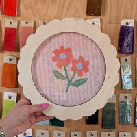 Hand holding a framed cross-stitched floral design on a wooden surface with color swatches.