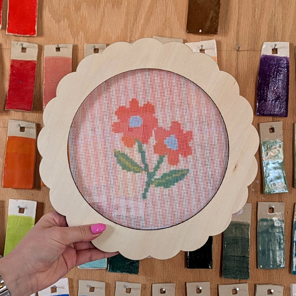 Hand holding a framed cross-stitched floral design on a wooden surface with color swatches.