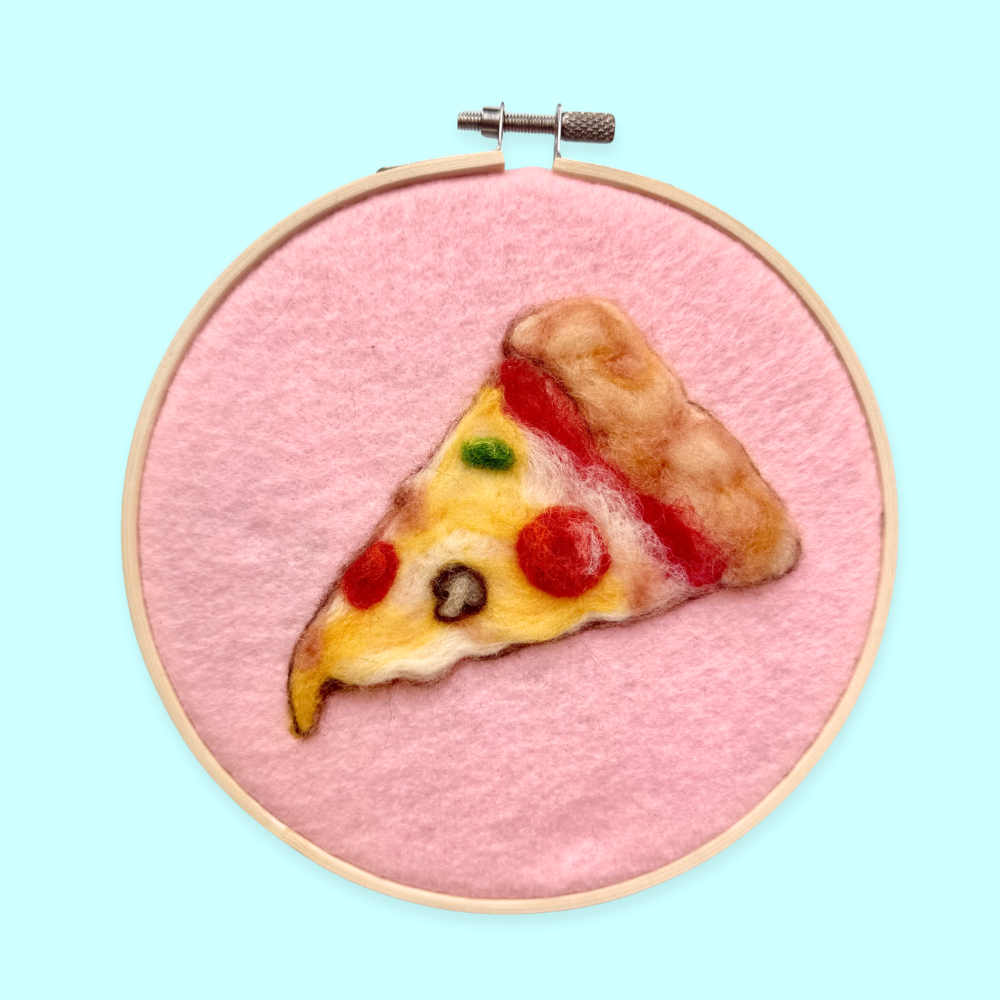 Embroidery hoop with a slice of pizza on pink fabric against a light blue background