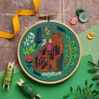 Embroidery hoop with a bear and floral design on a green fabric background, surrounded by embroidery tools and buttons.