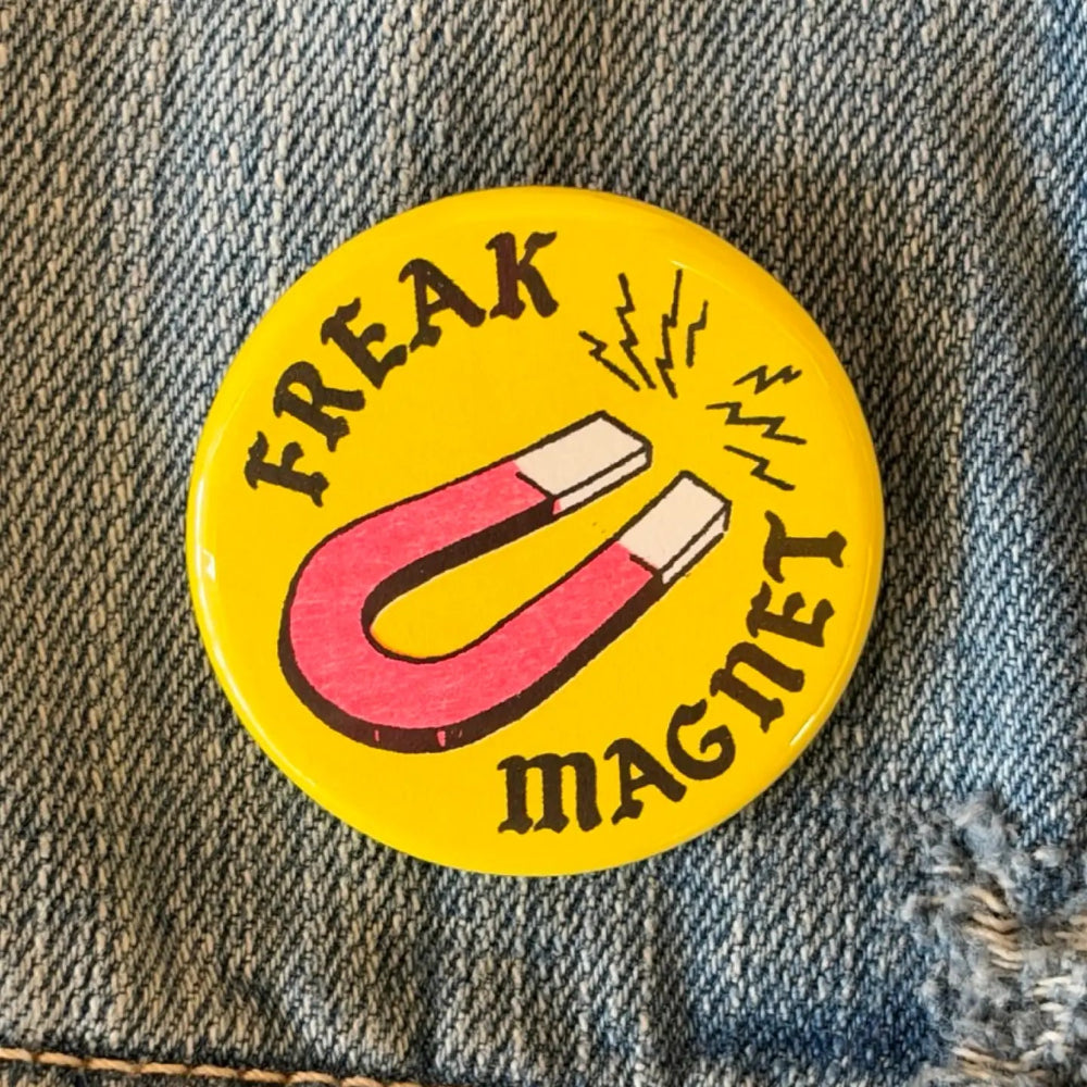 A yellow button against a denim background. The button has an illustration of a magnet in the middle with surrounding text that reads "Freak Magnet"