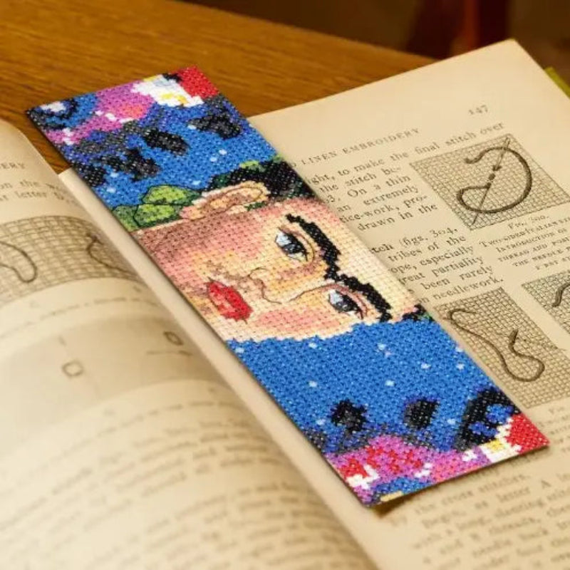 A colorful needlepoint-style bookmark featuring a stylized face and floral details, placed over an open book on a wooden surface