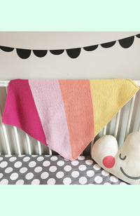 A knitted baby blanket with vertical stripes in pink, light pink, peach, and yellow, draped over a white crib next to a white cloud-shaped pillow.
