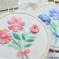 Embroidery hoops with floral designs on beige fabric, one in a variegated pink colorway and one in a variegated purple colorway. 