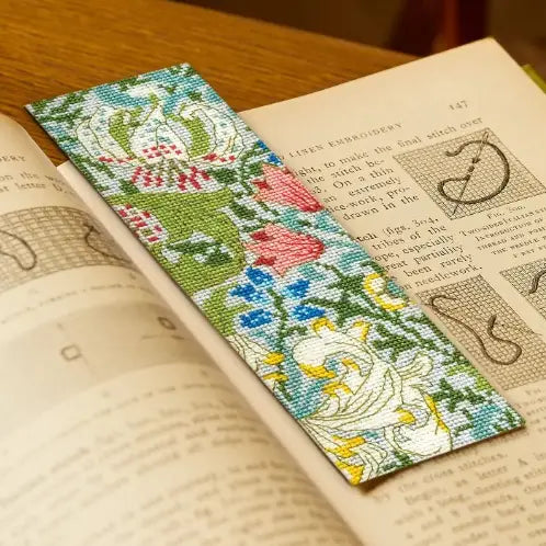 A rectangular cross-stitch style bookmark with a floral and bird pattern in green, blue, pink, yellow, and white, resting on an open book.