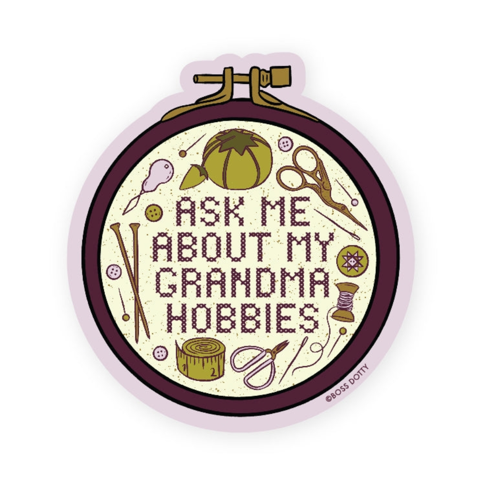 Text reads "Ask Me About My Grandma Hobbies" in cross-stitch within an embroidery hoop design in purple with various sewing and embroidery accessories in green and gold surrounding the text.