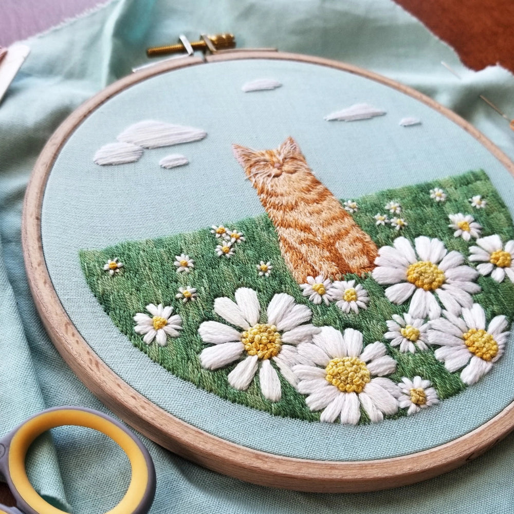 Embroidery hoop with a detailed design of an orange cat in a flower field