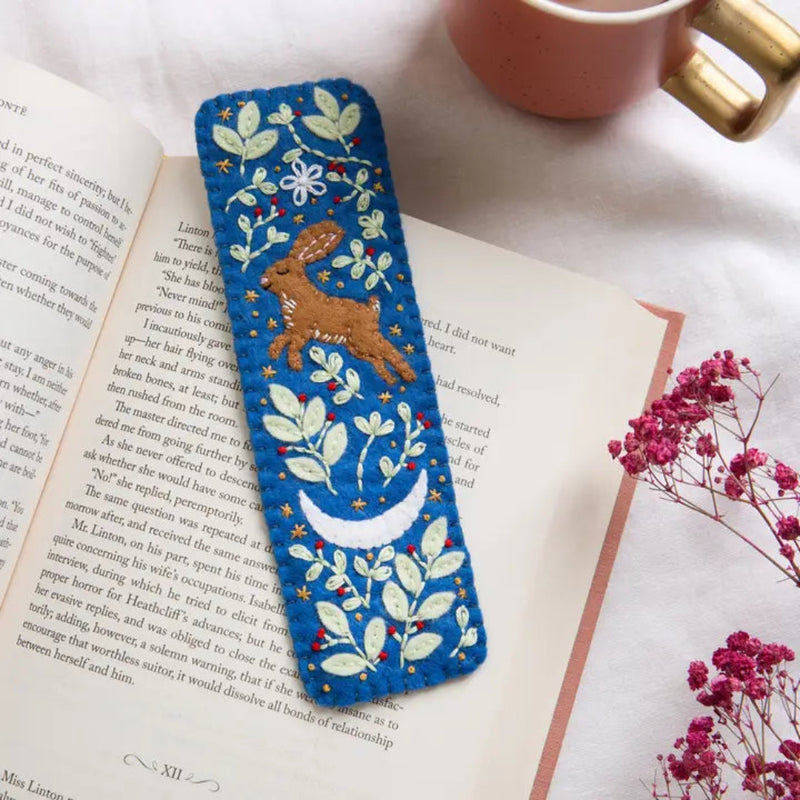 Embroidered bookmark with a hare design on an open book next to a cup and flowers.