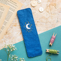 Blue embroidered bookmark with moon and stars on a textured surface with buttons and thread spools.