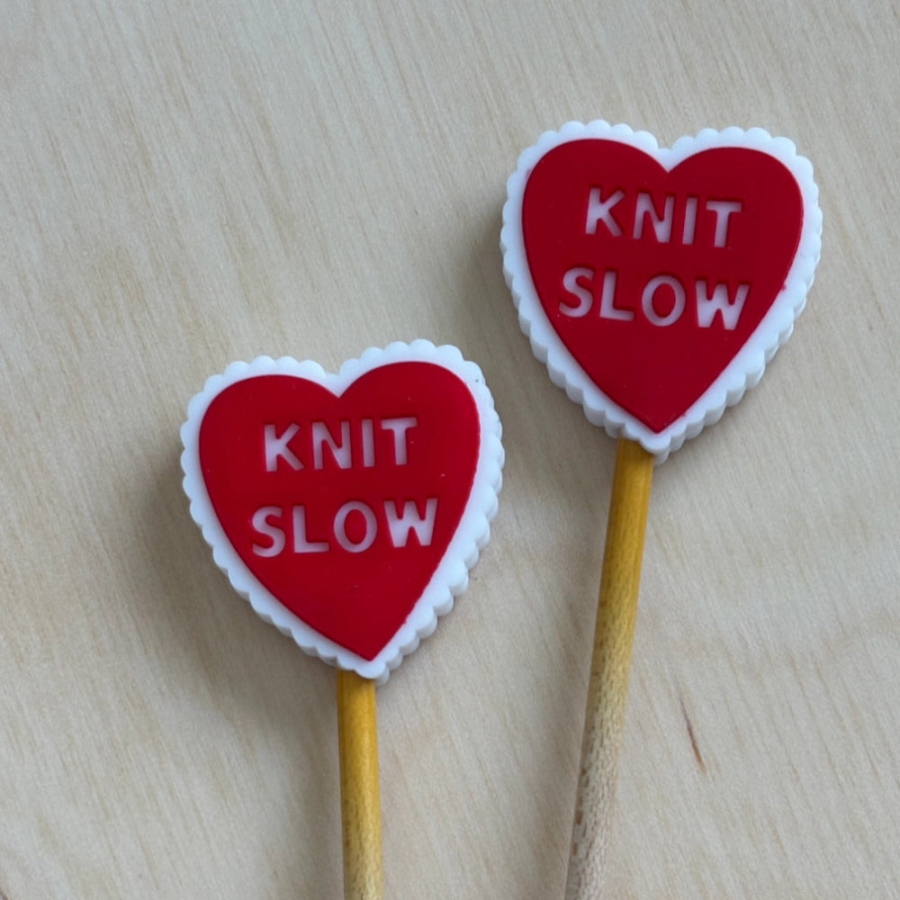 Two heart-shaped knitting needle tips with 'KNIT SLOW' text on a wooden surface.