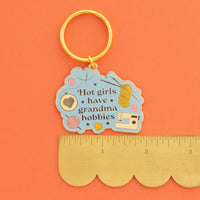 A novelty keychain with a gold-tone split ring and a light blue enamel charm featuring the text 'Hot girls have grandma hobbies' along with craft-themed illustrations, shown next to a gold-colored scalloped edge ruler on a solid orange background.