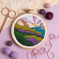 Embroidery hoop with a colorful landscape design on a pink surface with embroidery tools.