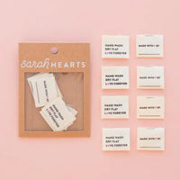 A set of fabric labels read "Hand Wash, Dry Flat, Love Forever" and "Made with ♥ by." are on a pink background. Some are also placed in packaging labeled "Sarah Hearts"