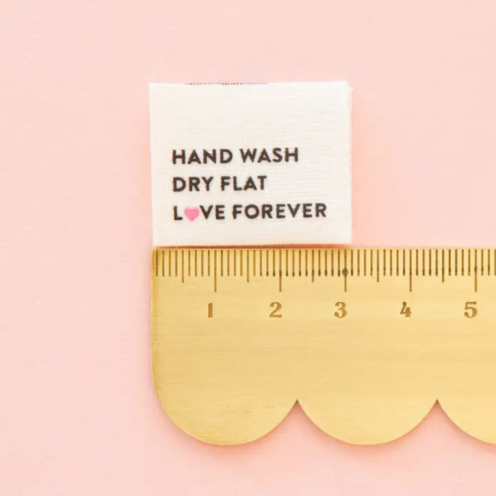 A fabric label reading "Hand Wash, Dry Flat, Love Forever" is on top of a scalloped edge ruler on a pink pink background. 