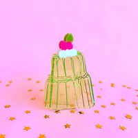 Clear green acetate hair claw in the shape of a two tier jello mold cake. Pink background with gold star confetti.