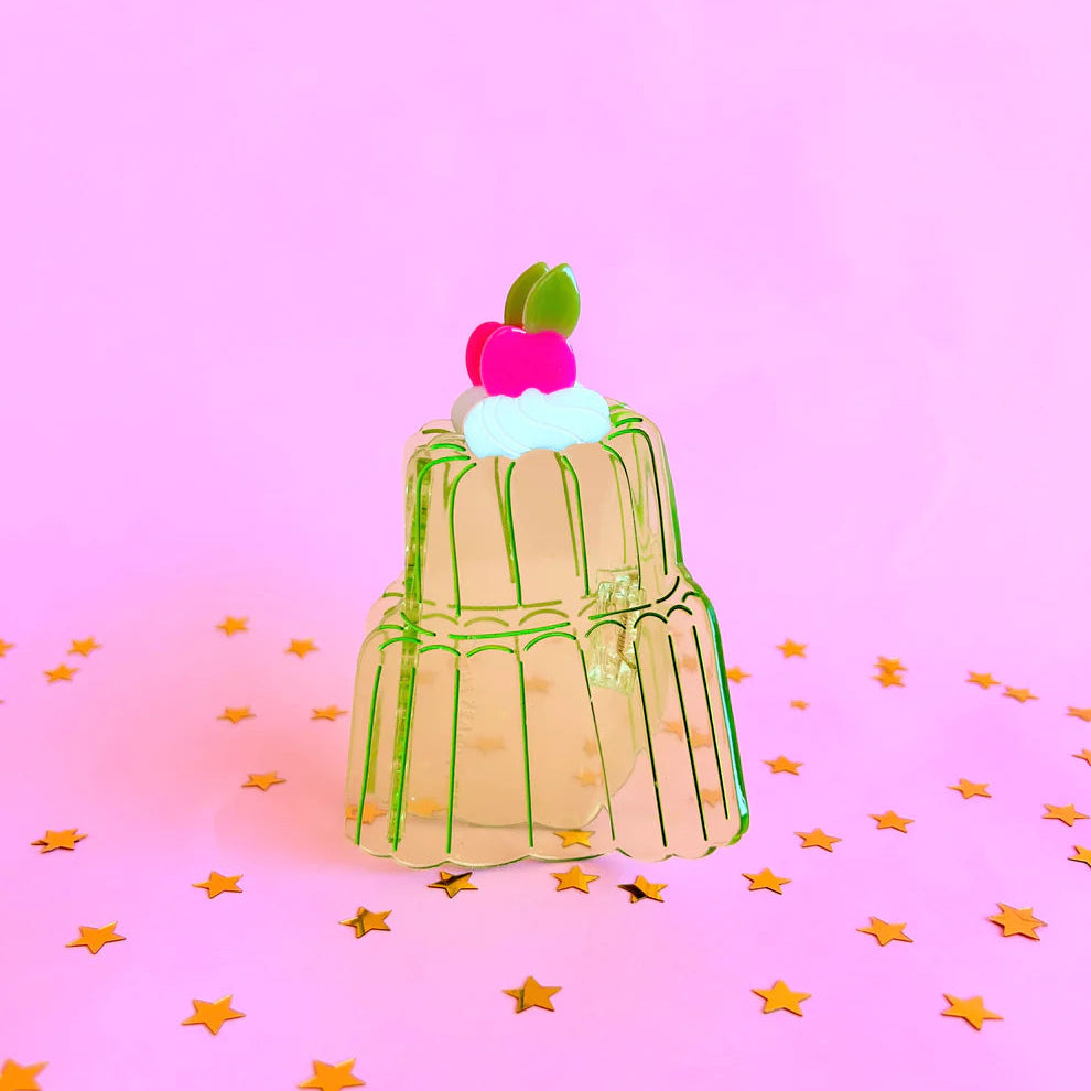 Clear green acetate hair claw in the shape of a two tier jello mold cake. Pink background with gold star confetti.
