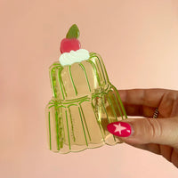 Clear green acetate hair claw in the shape of a two-tier jello mold with whipped cream and a cherry on top. Held in a hand with pink star nail art