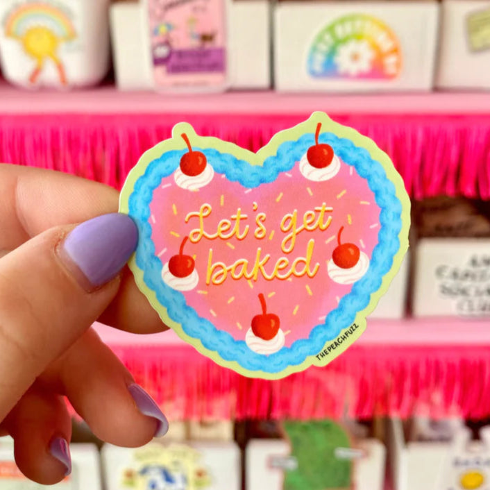 Hand with lilac nali polish holding a heart cake shapd sticker that reads "Let's get baked" in an frosting lettering style.