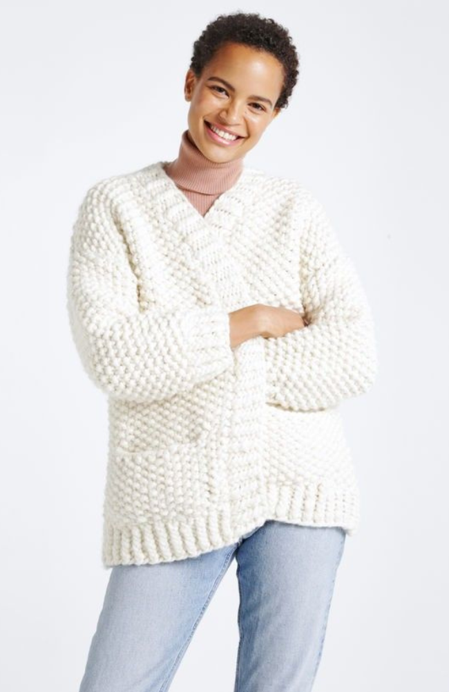 Woman with short brown curls dressed in light brown turtleneck, jeans, and a cream colored knit cardigan on a white background.