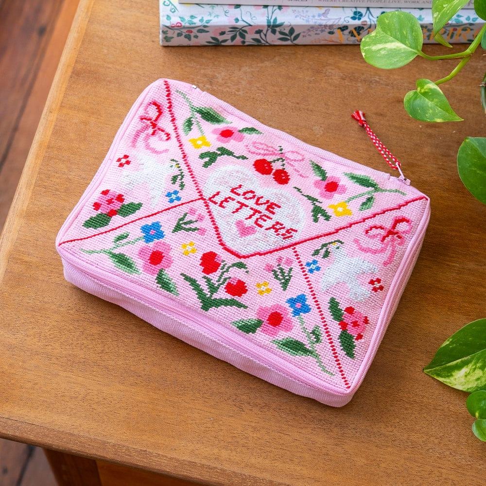 A rectangular zipper pouch with a needlepoint design resembling a pink envelope with the text 'love letters'