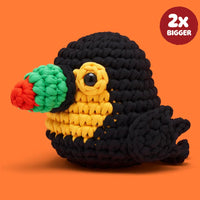 A crocheted plush toucan toy with a black body, yellow chest, green beak, and red beak tip, shown on an orange background with a red circular badge that reads '2x BIGGER'.